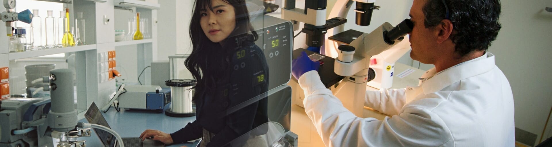 Woman on computer in lab and man looking into microscope